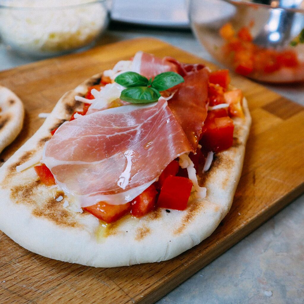 Focaccine veloci in padella su tagliere di legno, prosciutto crudo, pomodorini a cubetti, mozzarella e basilico fresco.